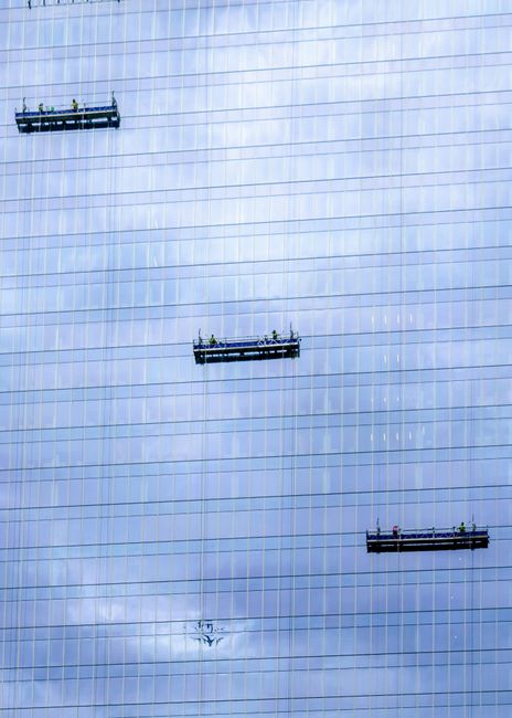Several window cleaners are working on the exterior of a tall, modern glass building, using suspended platforms to clean the reflective glass panels. The building's façade features numerous horizonta