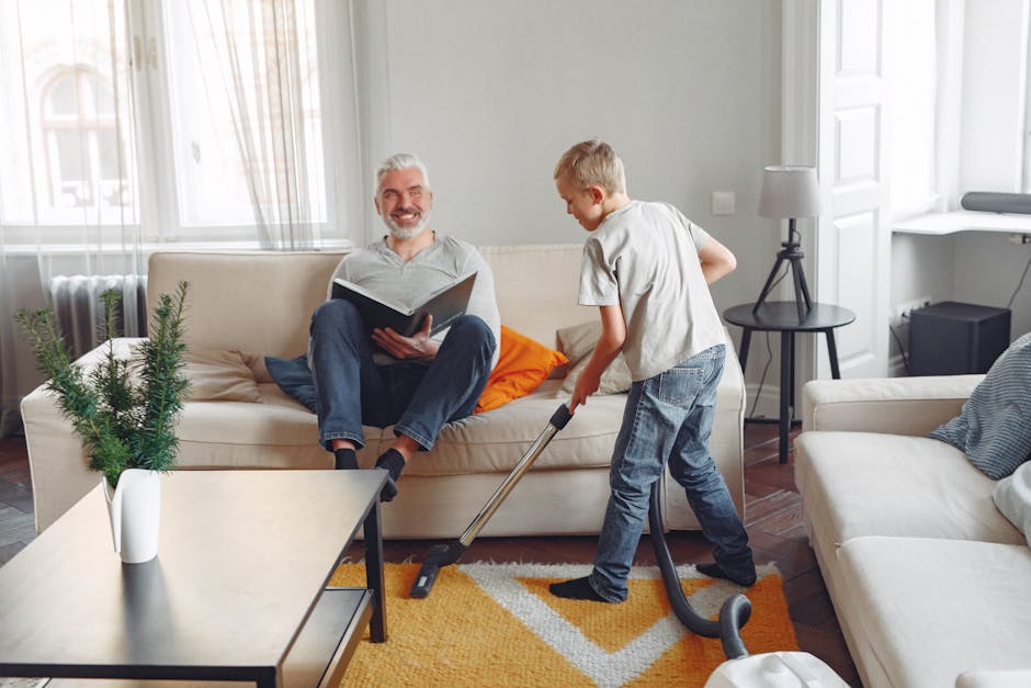 A man with grey hair and a beard is sitting on a beige sofa in a well-lit living room, smiling while holding an open magazine. A young boy, dressed in casual clothing, is vacuuming the orange and whit
