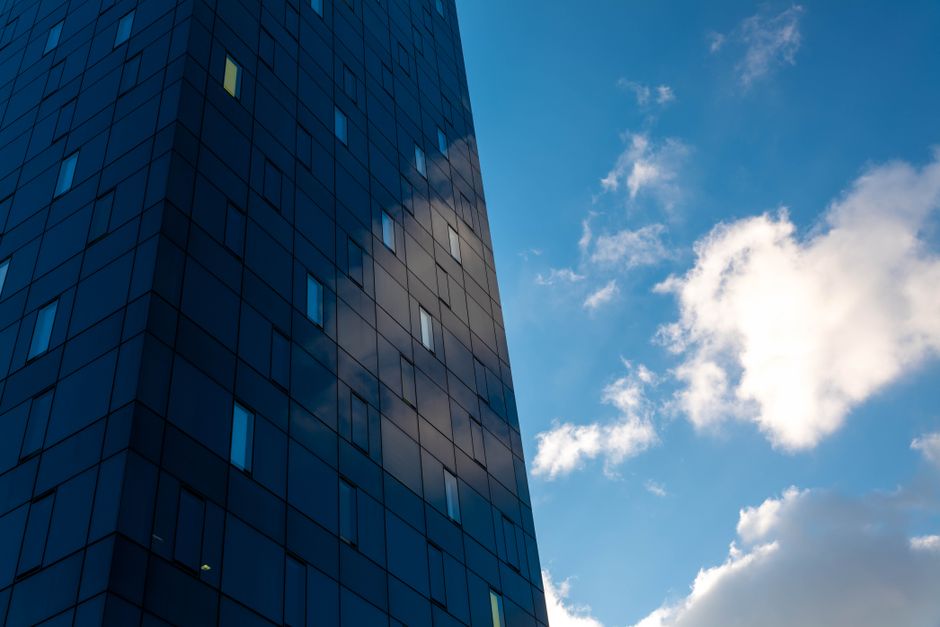 A group of four window cleaners wearing yellow safety helmets and harnesses are suspended on the glass facade of a tall modern office building. They are cleaning the large, reflective blue-tinted glas